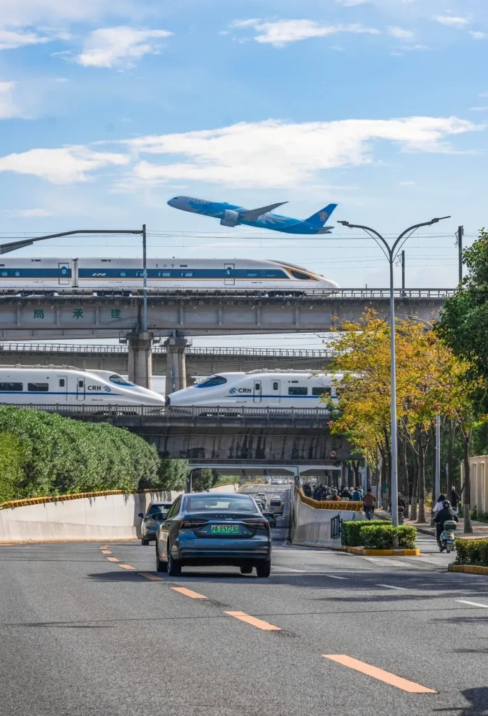 A blue and white airplane taking off in the sky above multiple levels of elevated high-speed trains and road traffic in a city.