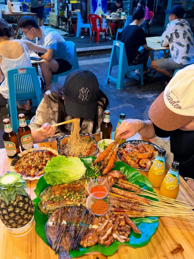 A wooden table laden with a variety of Dai foods, including a whole grilled fish on a large banana leaf, skewers of grilled shrimp and pork, grilled corn, a whole pineapple, bowls of dipping sauces, a plate of noodles, and bottles of beer.