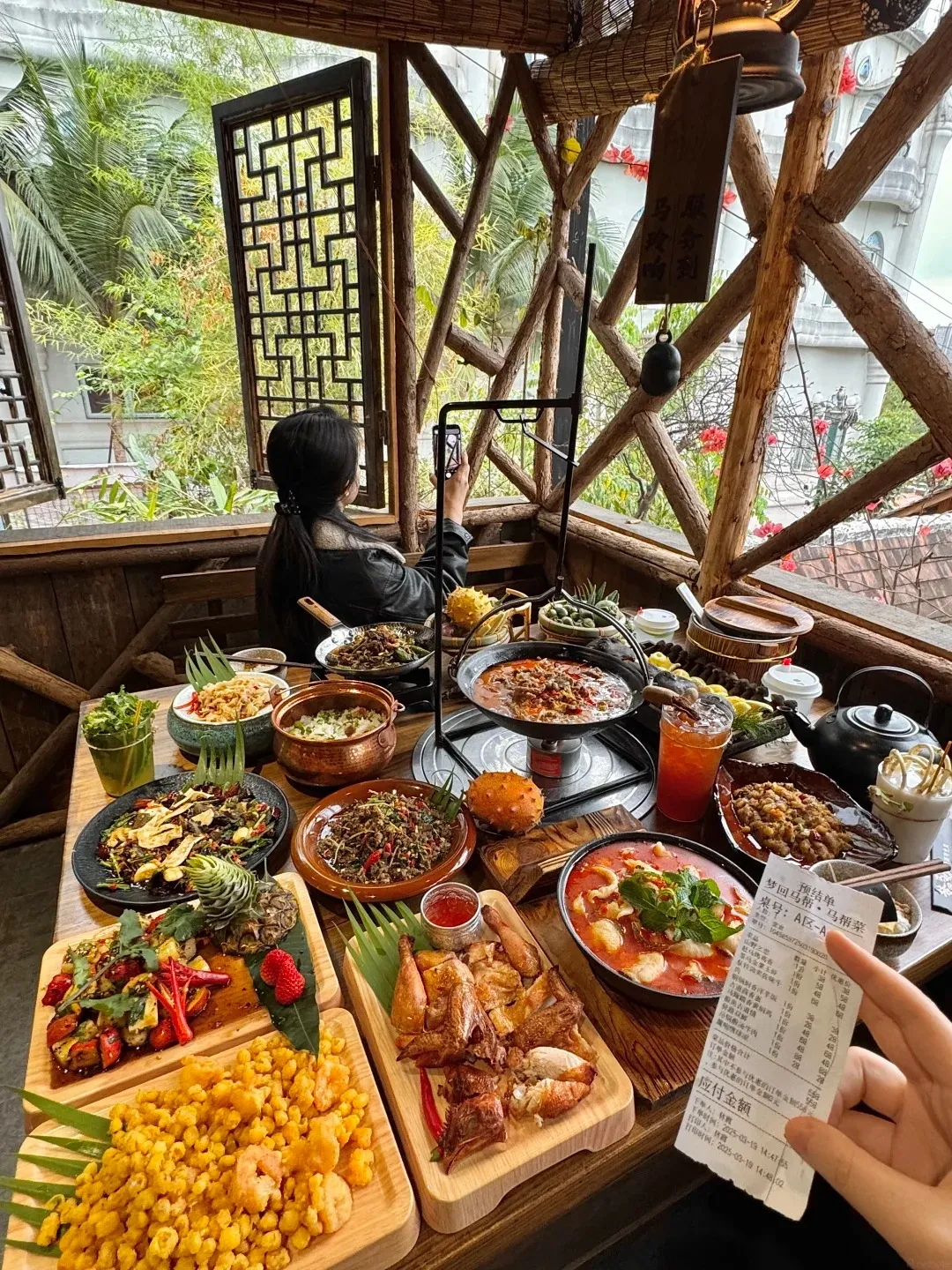 A table laden with various dishes of traditional Dai food, including roasted chicken, stir-fried vegetables, bowls of soup and rice, fried corn with shrimp, and a fruit resembling a spiky orange. A person is holding a receipt in the foreground.