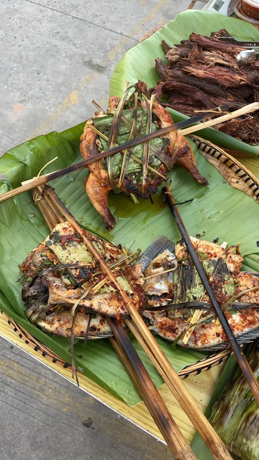 A whole grilled chicken wrapped in banana leaves and a grilled fish also wrapped in banana leaves, both served on a large banana leaf in a woven tray. A pile of dried meat is visible in the background.
