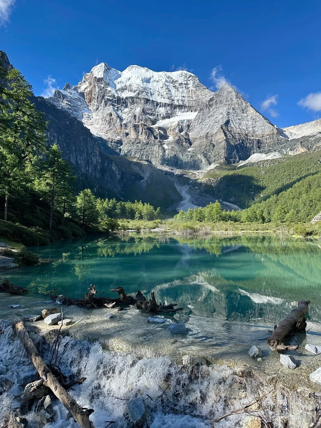 A turquoise lake with clear water reflecting snow-capped mountains and green trees under a blue sky. In the foreground, a small waterfall flows over rocks and fallen logs.