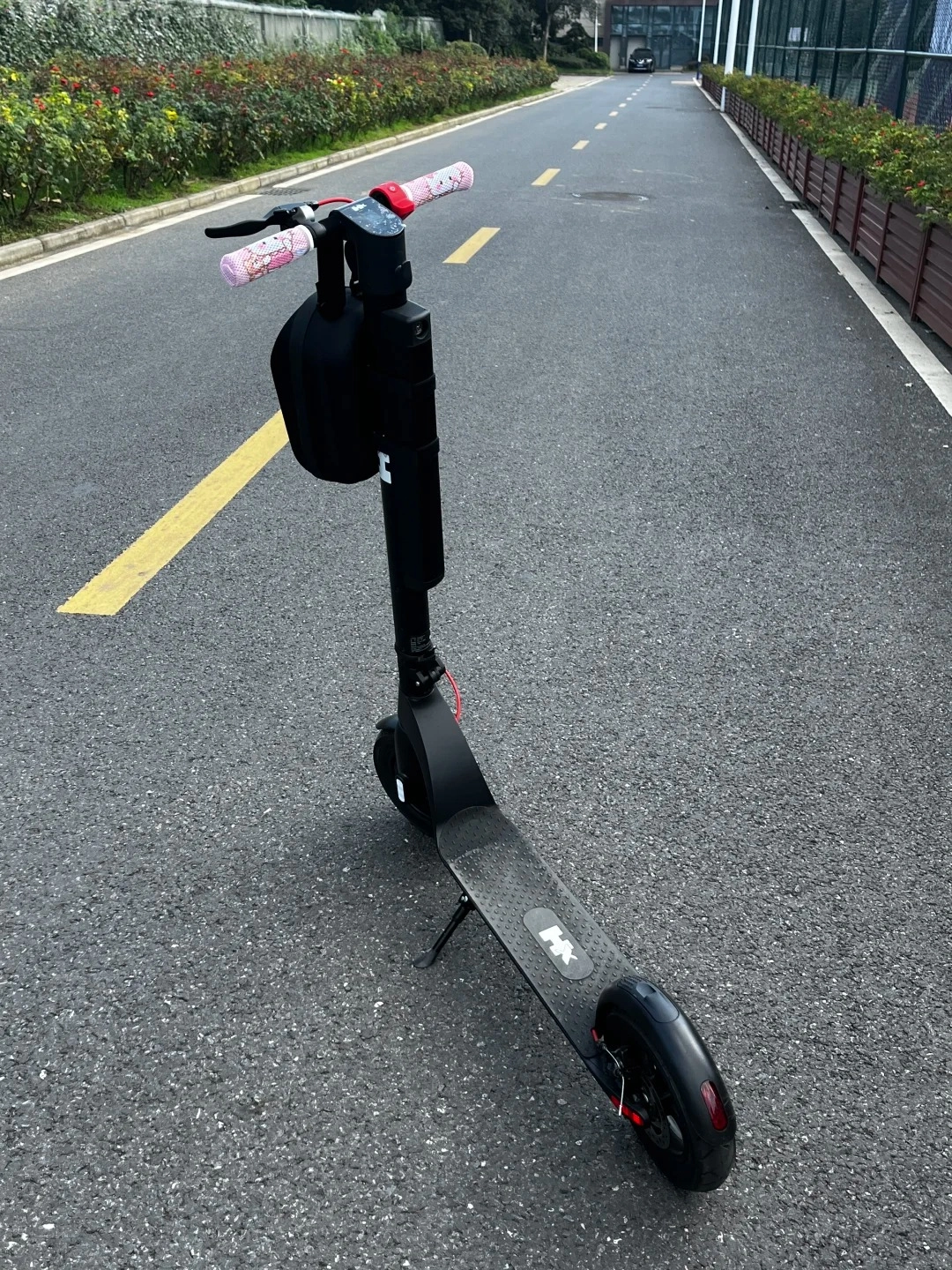 A black electric scooter with pink Hello Kitty patterned handlebar grips is parked on an asphalt road with yellow lane markings. The scooter has a black bag hanging from the handlebars and a kickstand deployed. The road is lined with bushes and a fence in the background.