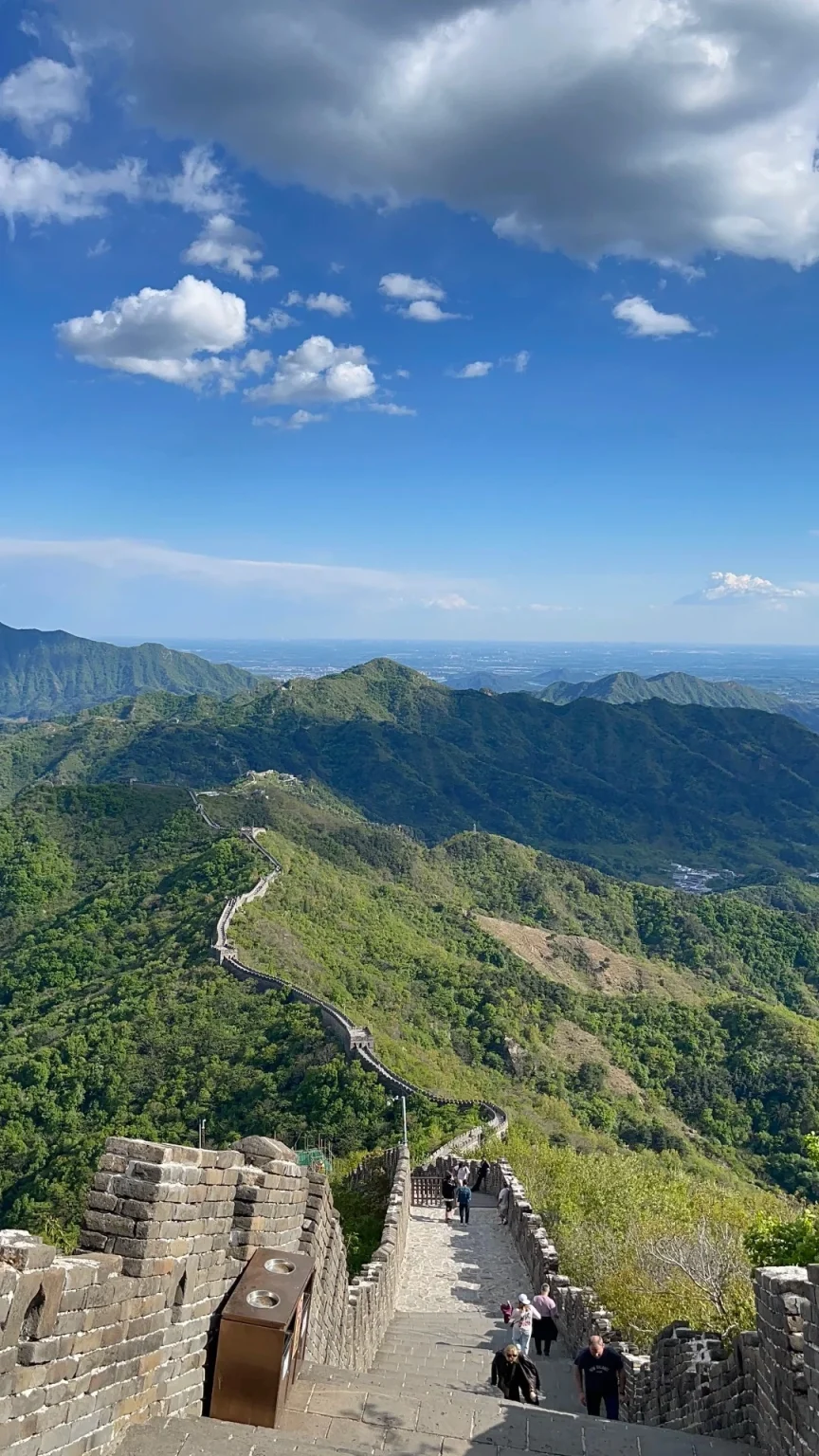 View from the Great Wall of China at Mutianyu, looking down a stone staircase lined with brick battlements. People are walking up and down the stairs. The wall continues in a winding path along lush green, tree-covered mountains under a bright blue sky with white and gray clouds.