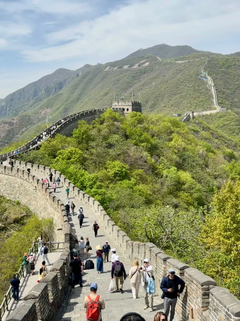 A wide view of the Great Wall of China winding over lush green, tree-covered mountains under a partly cloudy sky. Many tourists are walking along the stone pathway of the wall, which features watchtowers and crenelated battlements. In the foreground, the wall is wide with stone steps, and people are visible in casual clothing carrying bags and taking photos.