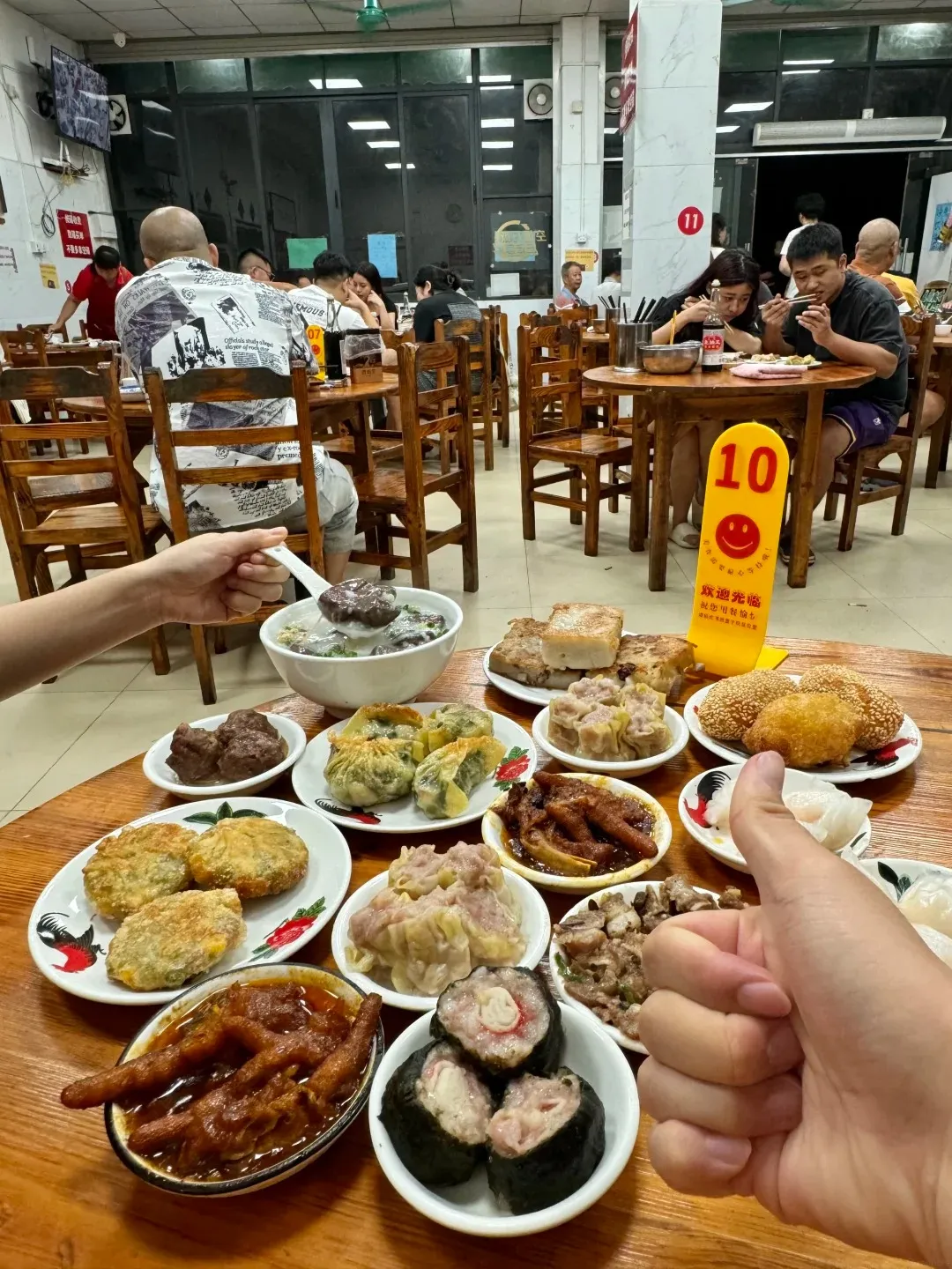 A close-up overhead view of a wooden table laden with various Chinese dim sum dishes, including steamed dumplings, fried items, chicken feet, and rolled sushi-like items. In the foreground, a hand gives a thumbs-up gesture. In the background, people are seated at tables in a busy restaurant.