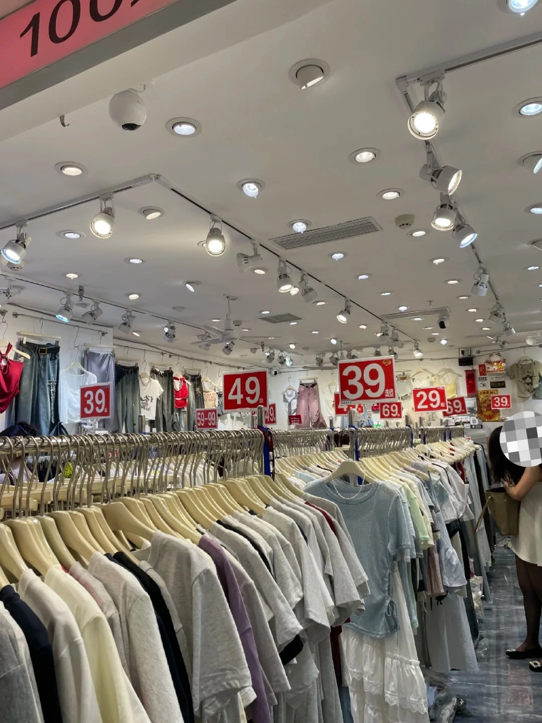 A view inside a clothing wholesale market in Guangzhou, with racks of t-shirts and other apparel on display, marked with red price tags showing prices like 39, 49, and 29.9. Several people are browsing the clothing, and spotlights illuminate the ceiling.