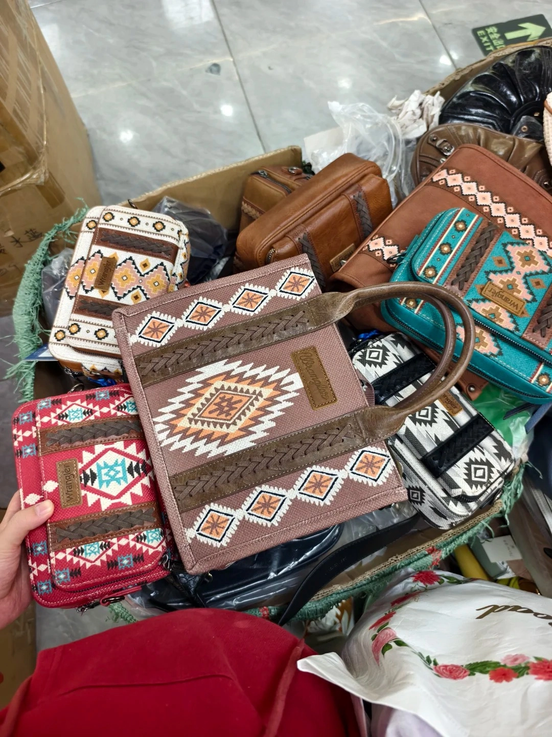 A collection of patterned handbags and clutches are piled in a box at a wholesale market. The bags feature Aztec-inspired geometric designs in brown, white, red, and teal. Some bags have braided leather accents and branded tags.