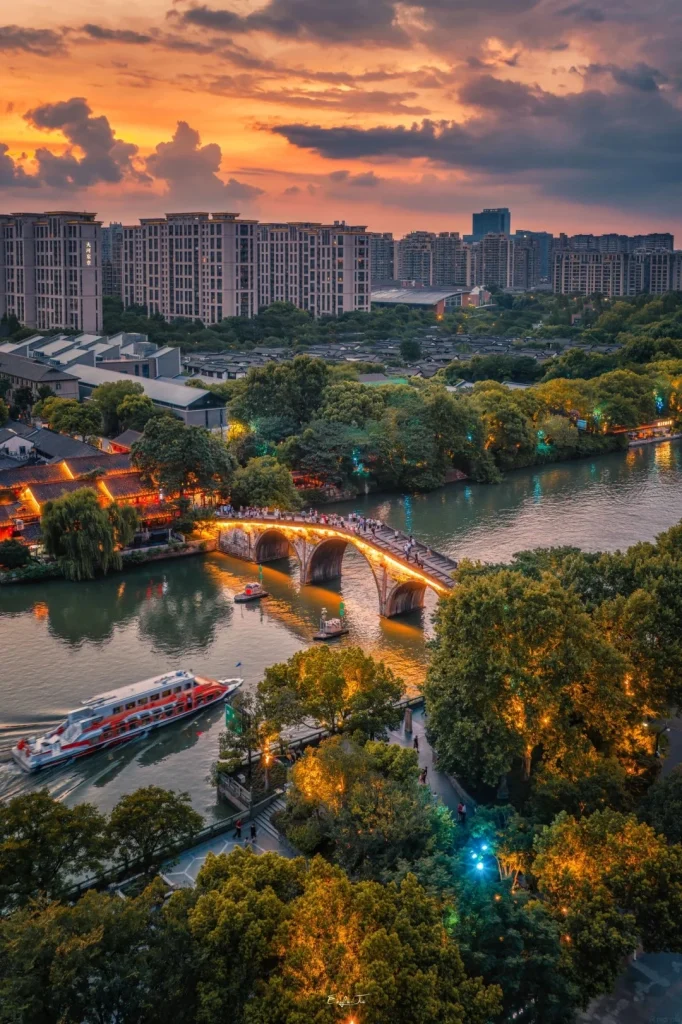 An aerial view of the Grand Canal in Hangzhou at sunset. A curved, stone bridge illuminated with lights spans the canal, with people walking across it. Several boats are on the water, including a long, red and white passenger ferry. The canal banks are lined with trees decorated with warm lights and traditional Chinese buildings with illuminated roofs. The sky above is filled with dramatic, colorful clouds in shades of orange, pink, and gray.