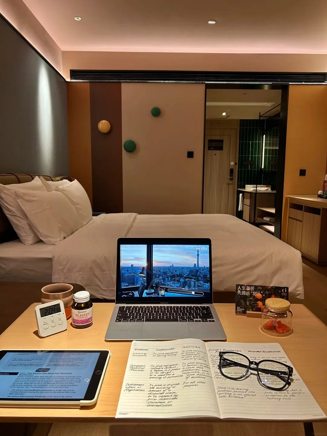 A wooden table in a hotel room with a laptop open, a tablet, a digital clock, a bottle of supplements, a notebook with handwritten notes and glasses, and a small jar of candies.