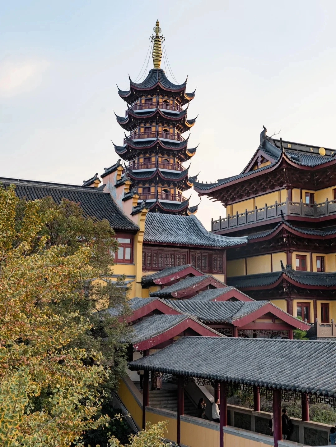 A multi-tiered traditional Chinese pagoda with dark curved roofs and red accents, rising above yellow and red temple buildings. In the foreground, a covered walkway with a dark tiled roof leads up stairs. Lush green and yellow foliage from trees is visible to the left.