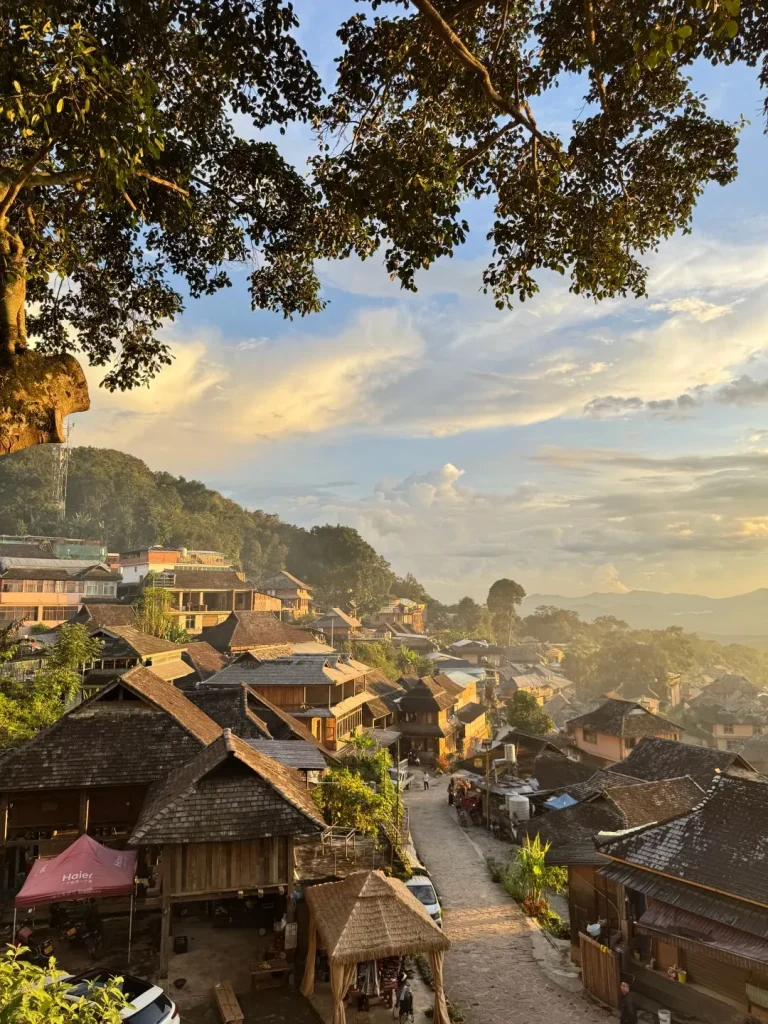A view of a traditional village nestled on a mountainside during sunset, with numerous wooden houses featuring sloped, shingled roofs. Trees with lush green foliage frame the top of the image, and the sky is filled with soft clouds illuminated by the warm glow of the setting sun. A paved road winds through the village, with a few people and vehicles visible.