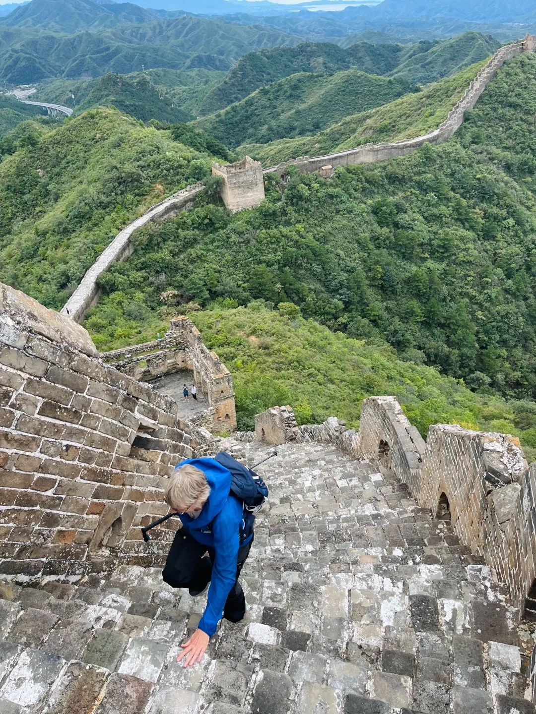 A person wearing a blue hooded jacket and a backpack is climbing up a steep, uneven stone staircase on the Great Wall of China. The person is looking down and reaching out with their left hand to steady themselves on the weathered bricks. In the background, the Great Wall winds along a lush green, mountainous landscape.
