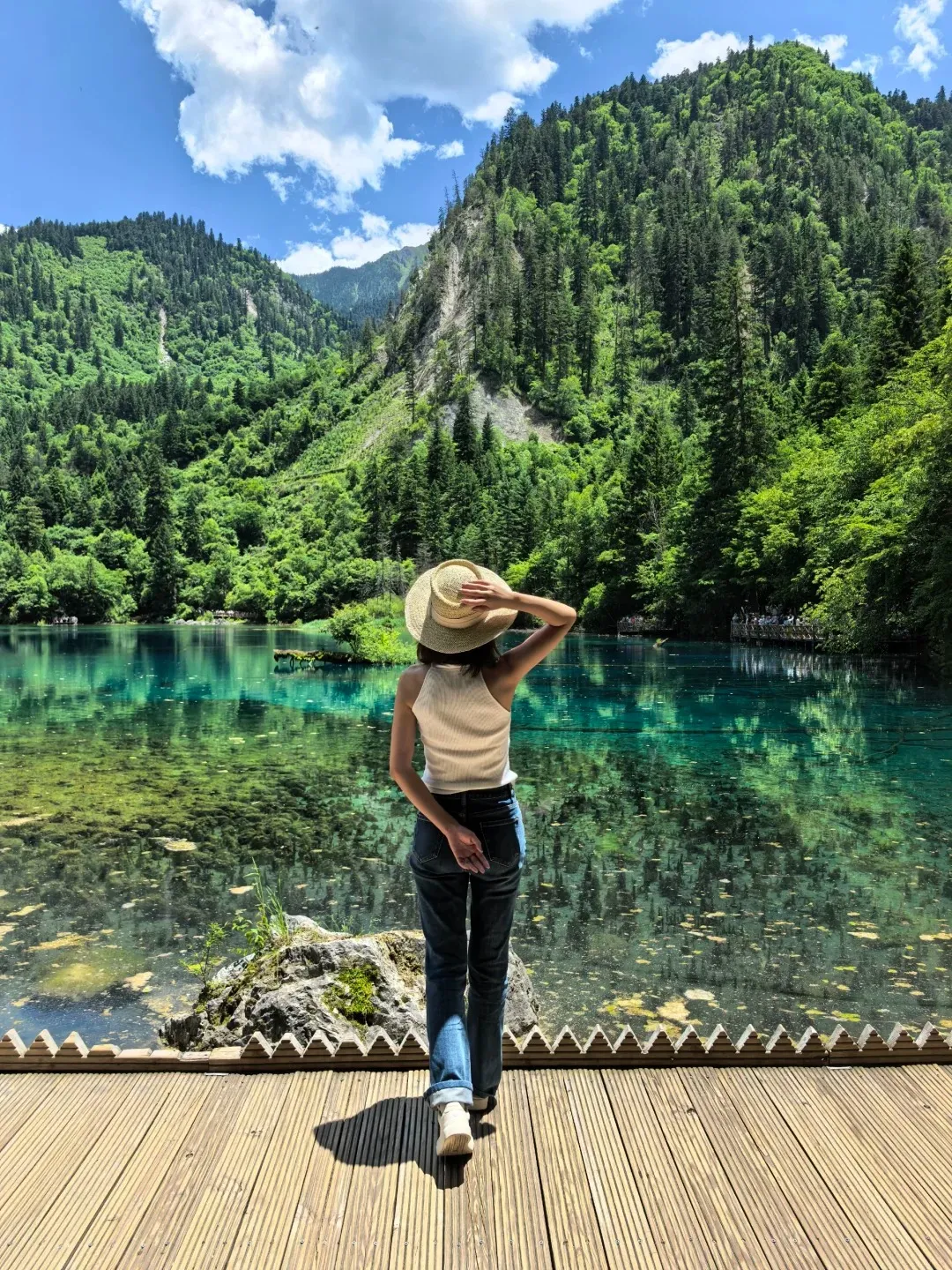 A person wearing a straw hat and jeans stands on a wooden boardwalk, facing away from the camera, looking out at the vibrant turquoise water of Five Flower Lake in Jiuzhaigou National Park. Lush green mountains covered in trees rise behind the lake under a blue sky with white clouds.