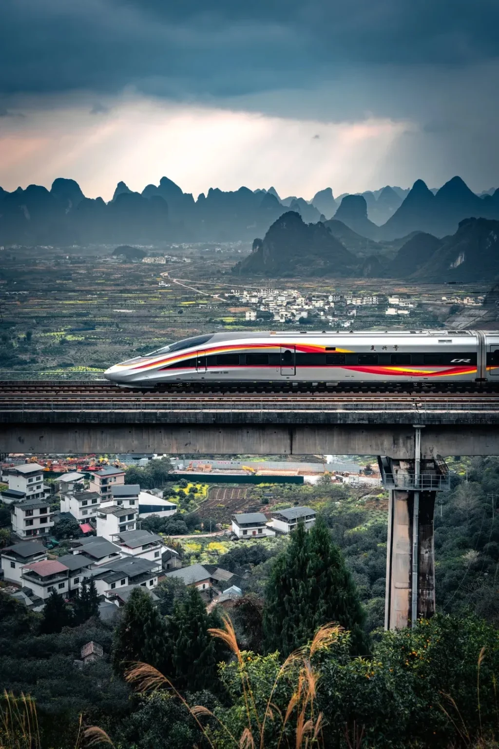A silver high-speed train with red and yellow stripes travels on an elevated railway bridge. Below the bridge, a village with white and dark-tiled houses is nestled amongst green trees and hills. In the background, a range of karst mountains under a dramatic cloudy sky stretches across the horizon.