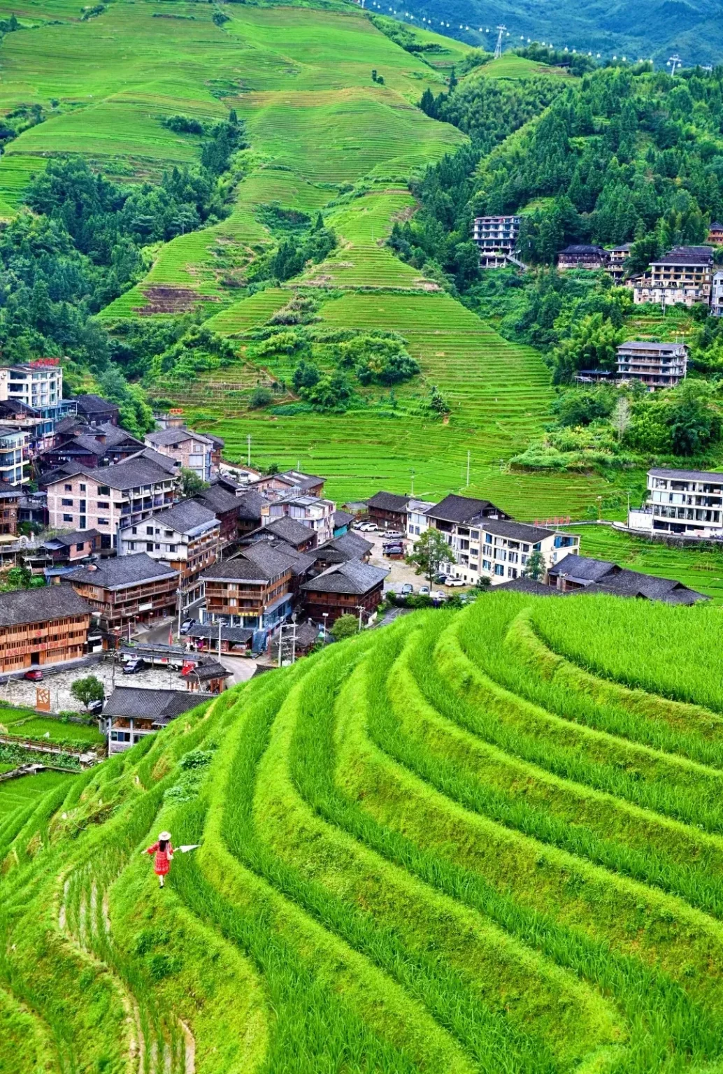 A person in a red dress with a straw hat walks on a narrow path through vibrant green, sculpted rice terraces that curve up a hillside. In the background, a village of traditional wooden and tiled buildings is nestled amidst the terraces and lush green trees, with more terraced hillsides and buildings visible further up the mountain.