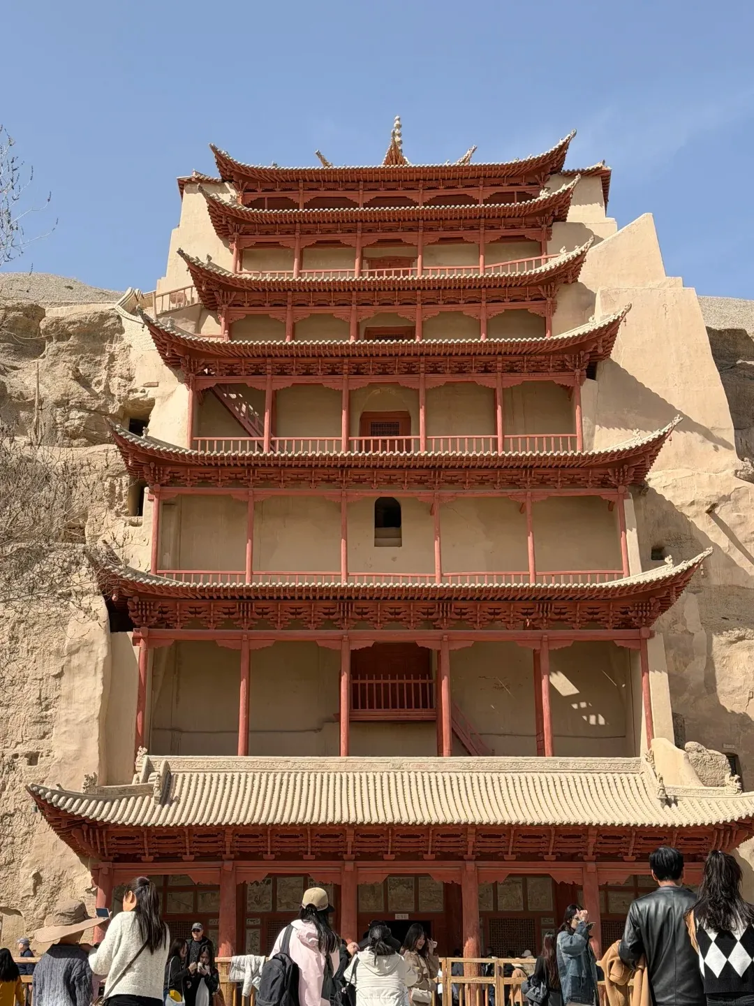 A tall, multi-tiered traditional Chinese wooden structure painted red, built into a sandstone cliff face. The building has numerous levels with curved roofs and balconies, and a large tiled roof overhang at the base. Several people are visible in the foreground.