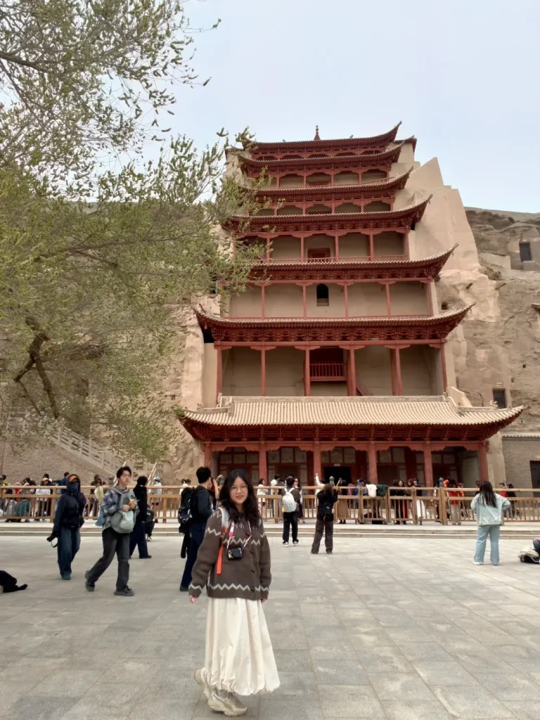 A young woman in a brown sweater and long cream skirt stands in a paved courtyard, smiling at the camera. Behind her, a large, multi-tiered traditional Chinese building with red painted eaves is built into a sandy cliff face. Several other tourists are visible in the background, some walking, some looking at the building.