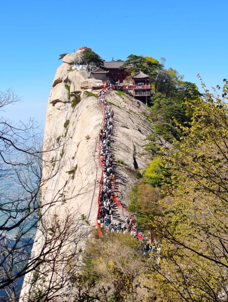 A steep, narrow stone staircase with a red railing winds up a sheer granite mountain face, crowded with a long line of hikers. Buildings, possibly temples, are visible at the summit of the mountain. Lush green trees and branches frame the lower right and left sides of the image, and a hazy landscape is visible in the distance.