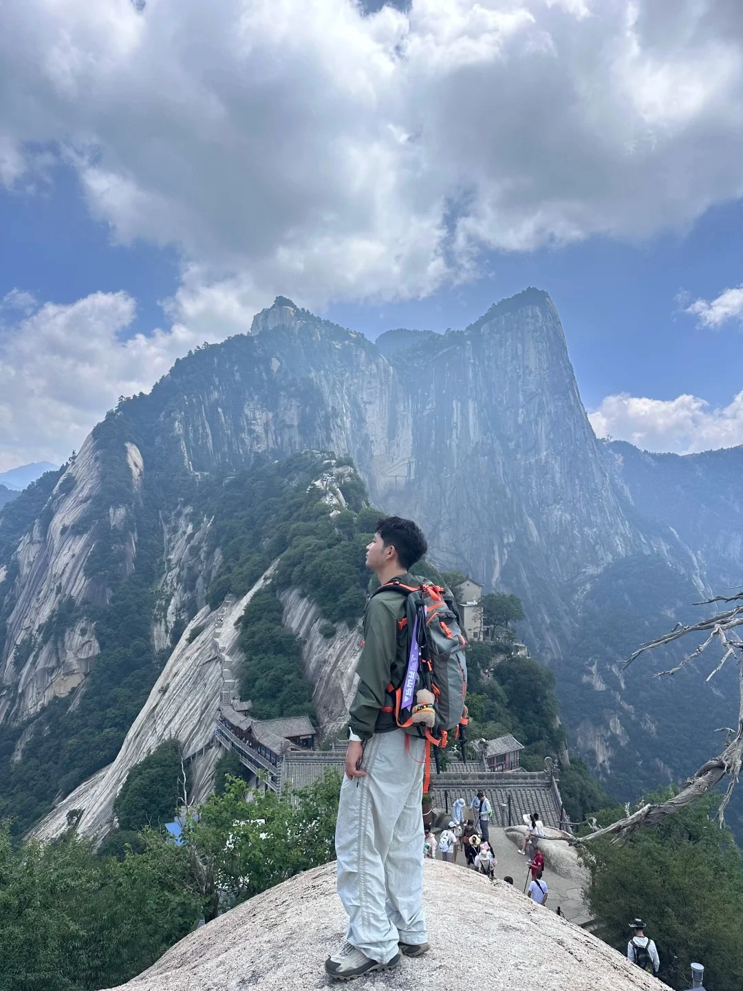 A young man with a backpack stands on a rocky peak, looking up at the majestic, steep mountains of Mount Huashan. In the background, a winding stone staircase with buildings and other tourists can be seen ascending the mountain.