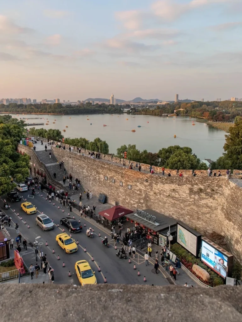 An elevated view looking down onto a wide, paved street running alongside a tall, ancient stone wall. Several yellow taxis and other vehicles are driving on the street, which is lined with pedestrians. A set of wide stairs leads up to the top of the wall where more people are walking. Beyond the wall, a large lake is visible with numerous small boats on it, and a city skyline in the distance under a hazy, pale sky.