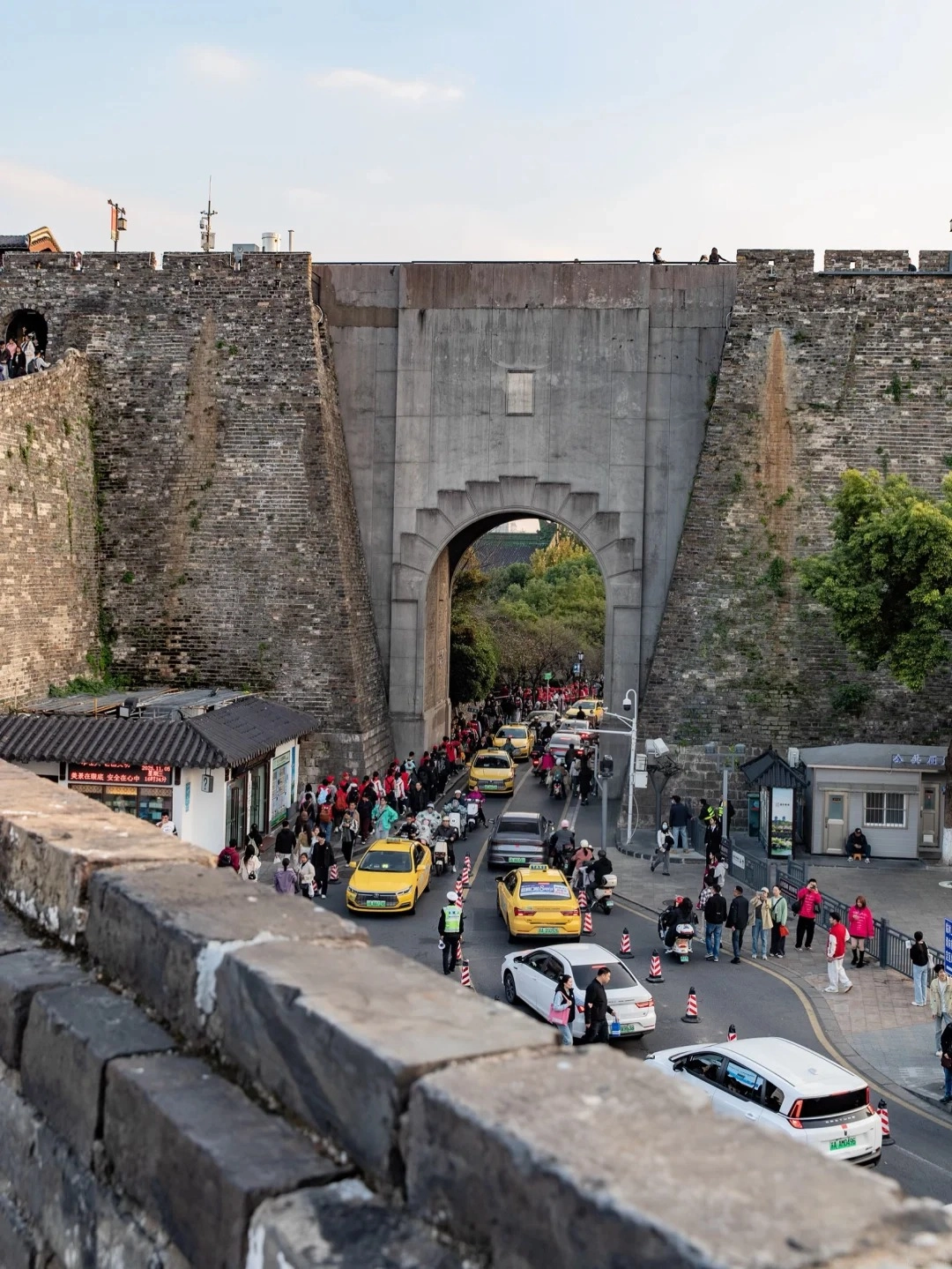 View from atop a weathered brick city wall looking down at a wide street leading through a large, arched stone gateway. Yellow taxis, white cars, motorcycles, and a crowd of people are on the street. The gateway is part of a massive, ancient stone fortification with some modern concrete additions.