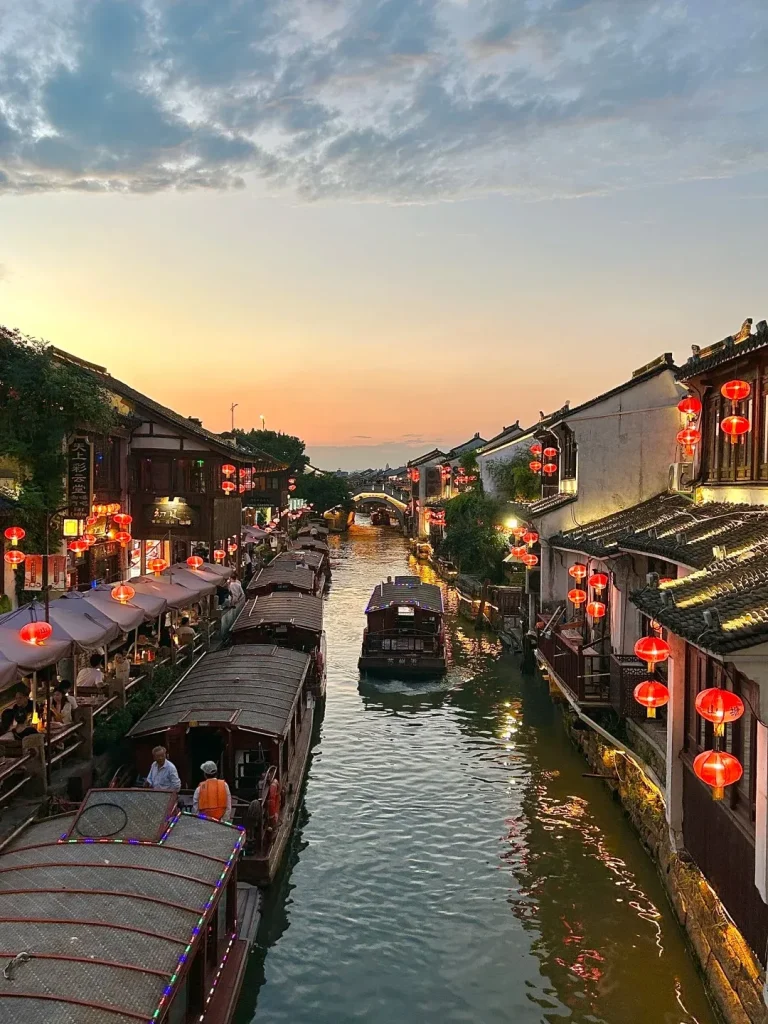 A scenic view from above a canal in Suzhou, China, at sunset. Traditional buildings with tiled roofs line the canal, adorned with glowing red lanterns. Several boats are on the water, some with people on board, and others are docked along the side where there are shops and eateries. The sky transitions from a soft orange and yellow near the horizon to a pale blue with scattered clouds.