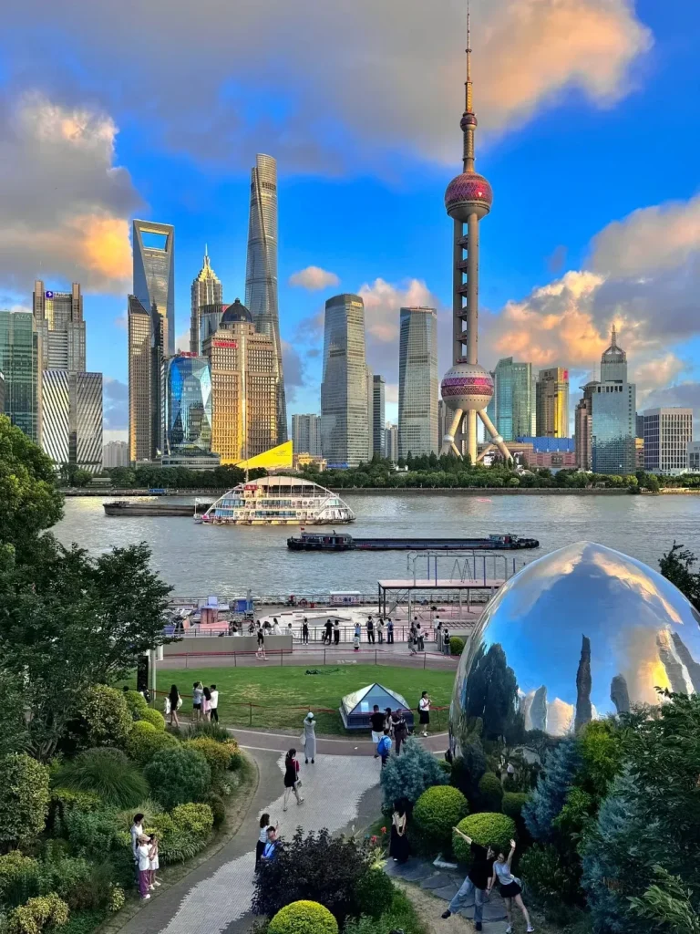 A panoramic view of the Shanghai skyline across the Huangpu River, featuring numerous skyscrapers and the Oriental Pearl Tower. In the foreground is a lush green park with people walking and a large, reflective spherical sculpture.