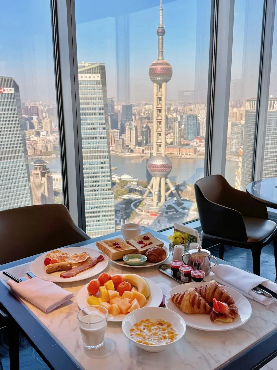 A luxurious hotel breakfast spread on a marble table with a panoramic view of Shanghai's skyline, including the Oriental Pearl TV Tower.