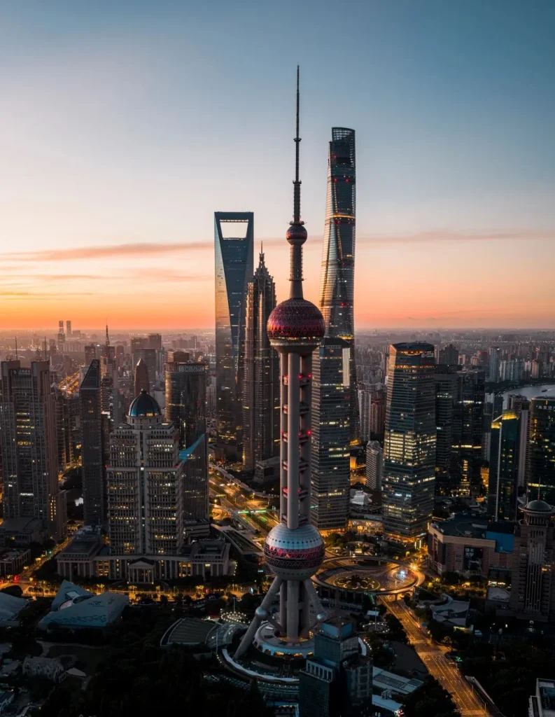 An aerial, panoramic view of the Shanghai Lujiazui skyline at dusk. Prominently featured in the foreground is the Oriental Pearl Tower, a distinctive TV tower with spherical elements. To its right is the Shanghai Tower, a modern supertall skyscraper. To the left of the Oriental Pearl Tower is the Jin Mao Tower, another skyscraper. Surrounding these landmarks are numerous other illuminated high-rise buildings, casting reflections on the faintly visible streets and infrastructure below. The sky above transitions from a warm orange and pink sunset glow near the horizon to a clear blue at the top.