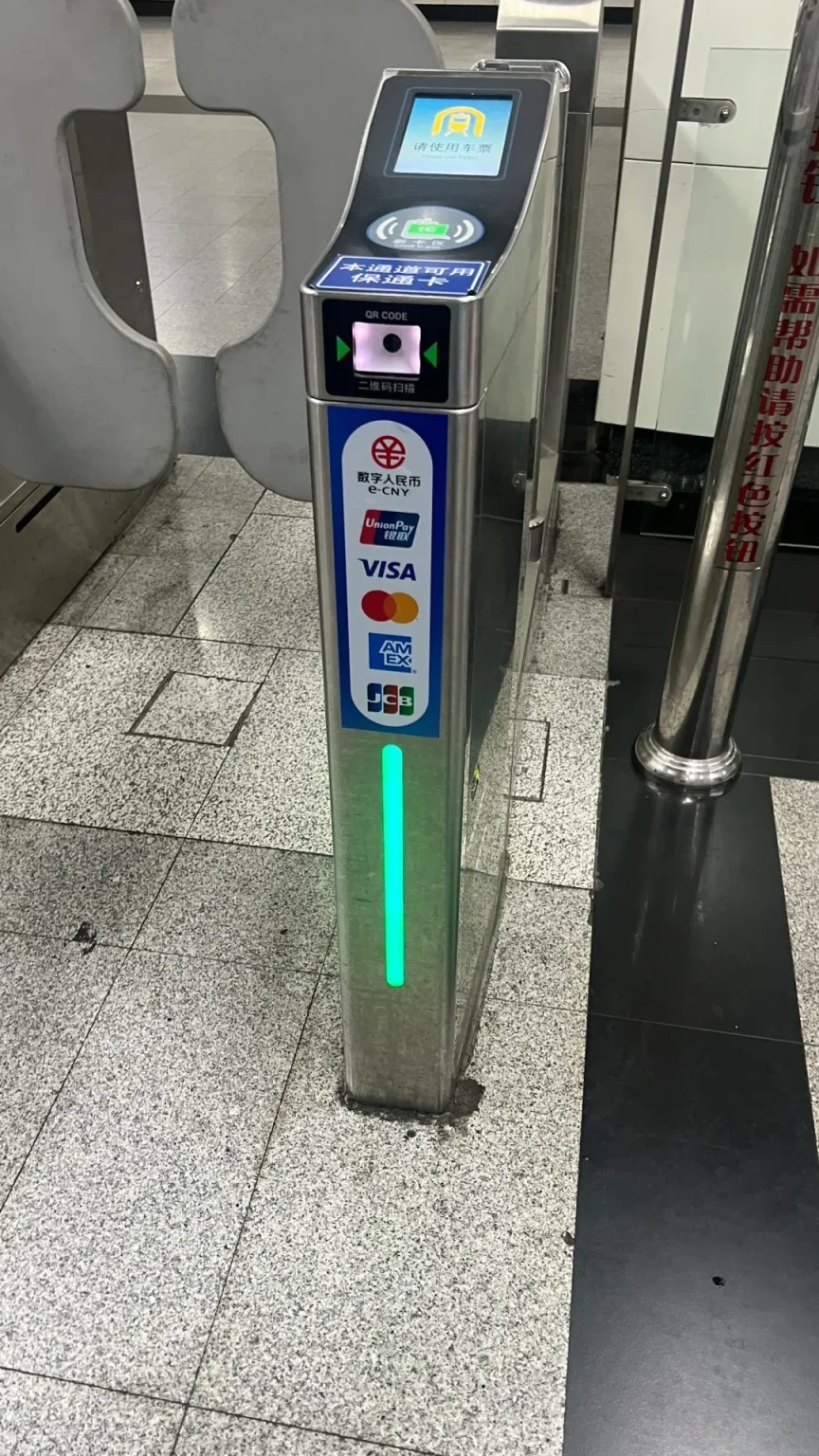 A close-up of a turnstile gate at a Shanghai Metro station. A person is holding a smartphone with a QR code displayed, about to scan it at the designated reader. The turnstile has various payment method logos including UnionPay, Visa, AMEX, and JCB, as well as Chinese characters indicating QR code scanning and different payment options.