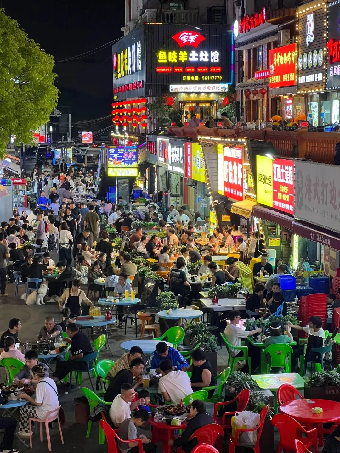 A bustling night market street in Shanghai, packed with people dining at numerous outdoor tables under bright neon signs and red lanterns. Food stalls are visible with cooking activities, and the atmosphere is lively and crowded.