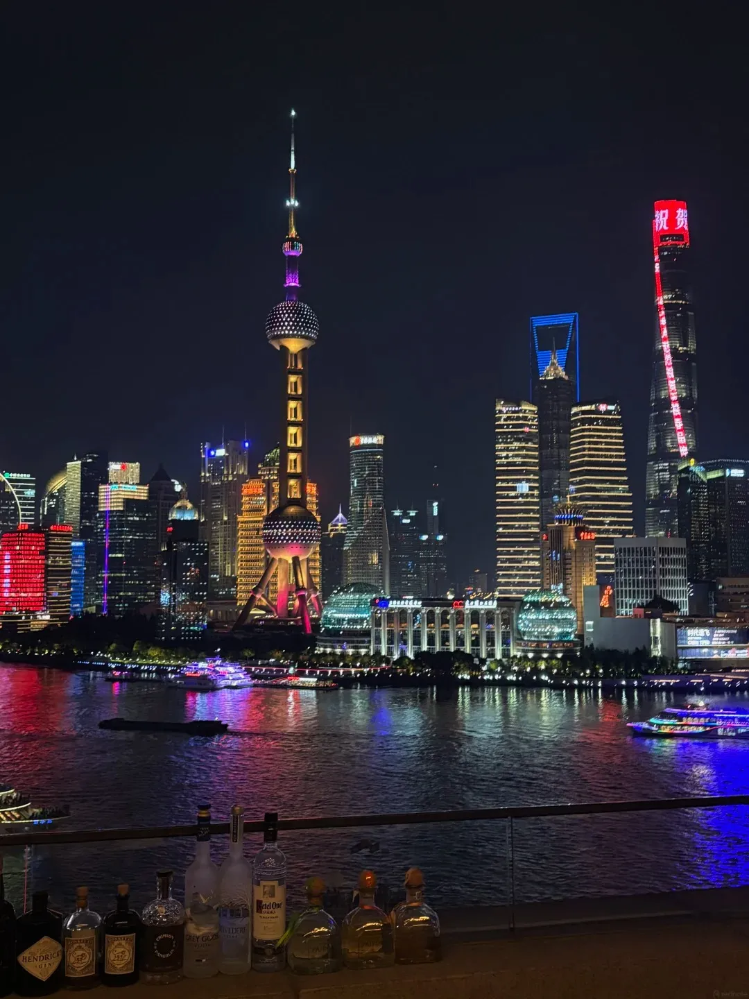 A nighttime view of the Shanghai skyline across a body of water, with illuminated skyscrapers and the Oriental Pearl Tower. In the foreground, a row of liquor bottles sits on a ledge.