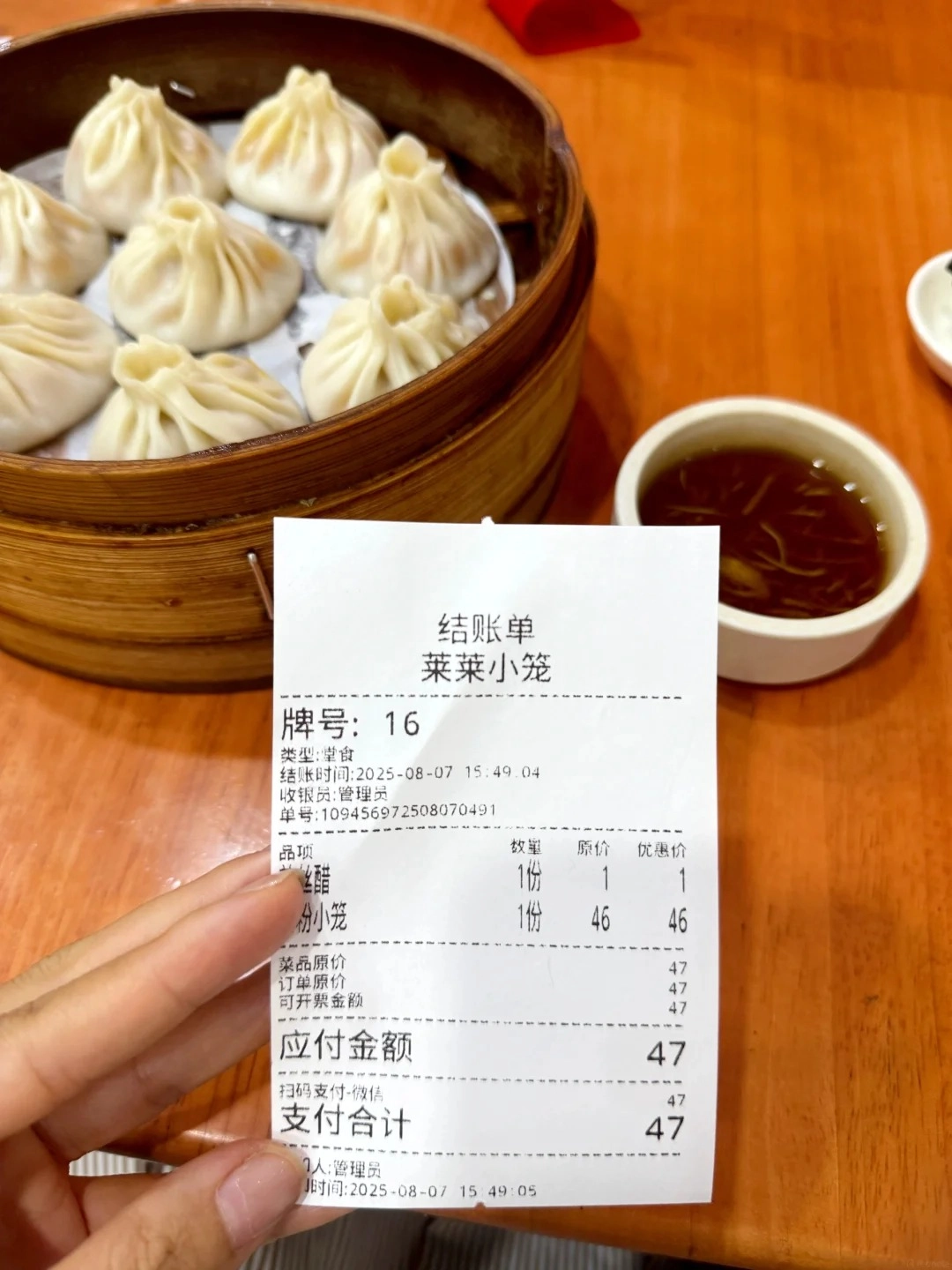 A receipt for xiaolongbao is held by a hand in front of a bamboo steamer basket filled with seven soup dumplings. A small bowl of dipping sauce is to the right.