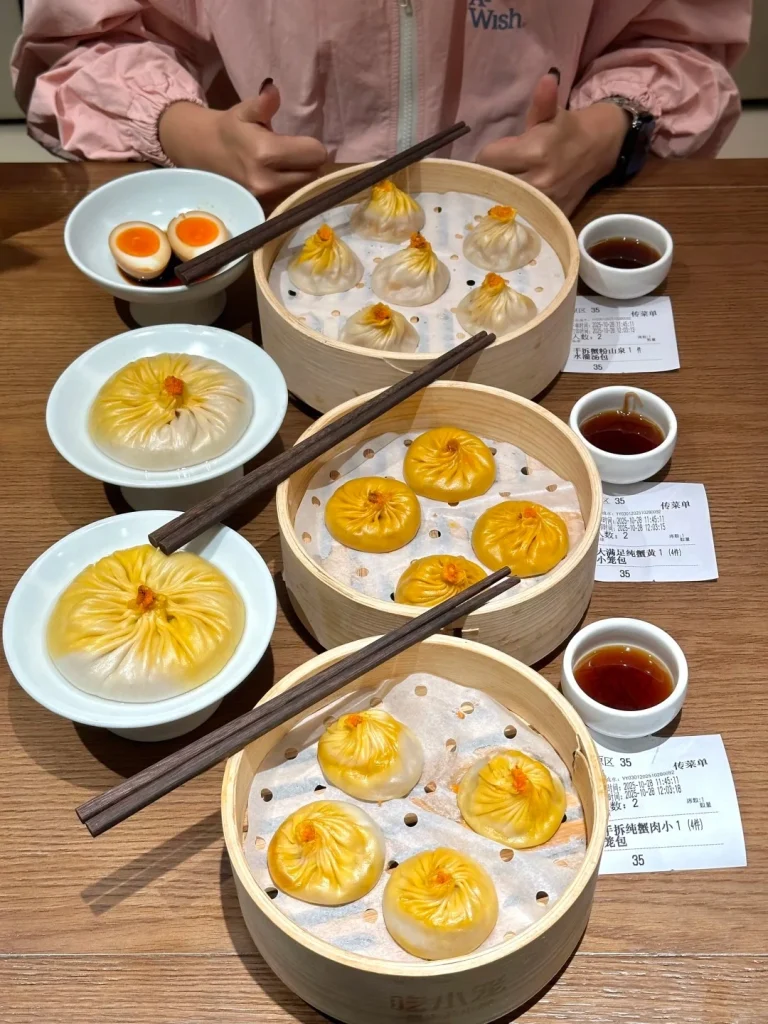 Overhead view of a wooden table set with several bamboo steamers filled with Shanghai Xiaolongbao soup dumplings. One steamer in the foreground contains five dumplings topped with orange roe. Another steamer behind it holds six dumplings. To the left, two white bowls are visible: one with two halved soft-boiled eggs in soy sauce, and another with a large, golden-yellow soup dumpling. Small white ceramic cups filled with dark dipping sauce are placed near the steamers. Chopsticks rest across the steamers and on the table. A person's hands, wearing a pink jacket, are visible at the top of the frame, giving a thumbs up. Receipts with Chinese text are placed near the dipping sauce cups.