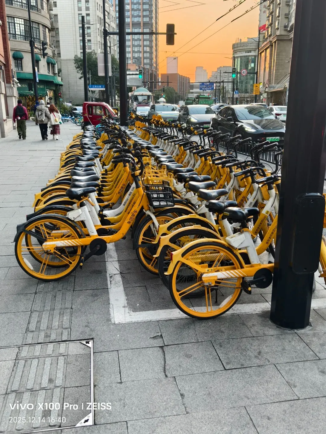 A row of bright yellow shared bicycles parked on a city sidewalk in Shanghai. The handlebars and wheels of the bikes are visible, with some showing Chinese characters on their frames. In the background, there are buildings, traffic lights, cars, and people walking on the sidewalk.