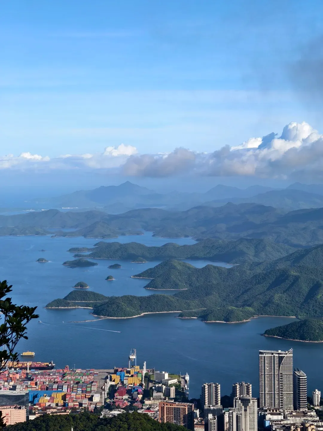 A panoramic view from a high vantage point overlooking a bay filled with numerous green, tree-covered islands. In the foreground, a bustling container port with stacks of colorful shipping containers and cranes is visible, along with several modern high-rise buildings and some foliage in the lower left corner. The sky is blue with scattered white clouds and distant misty mountains.