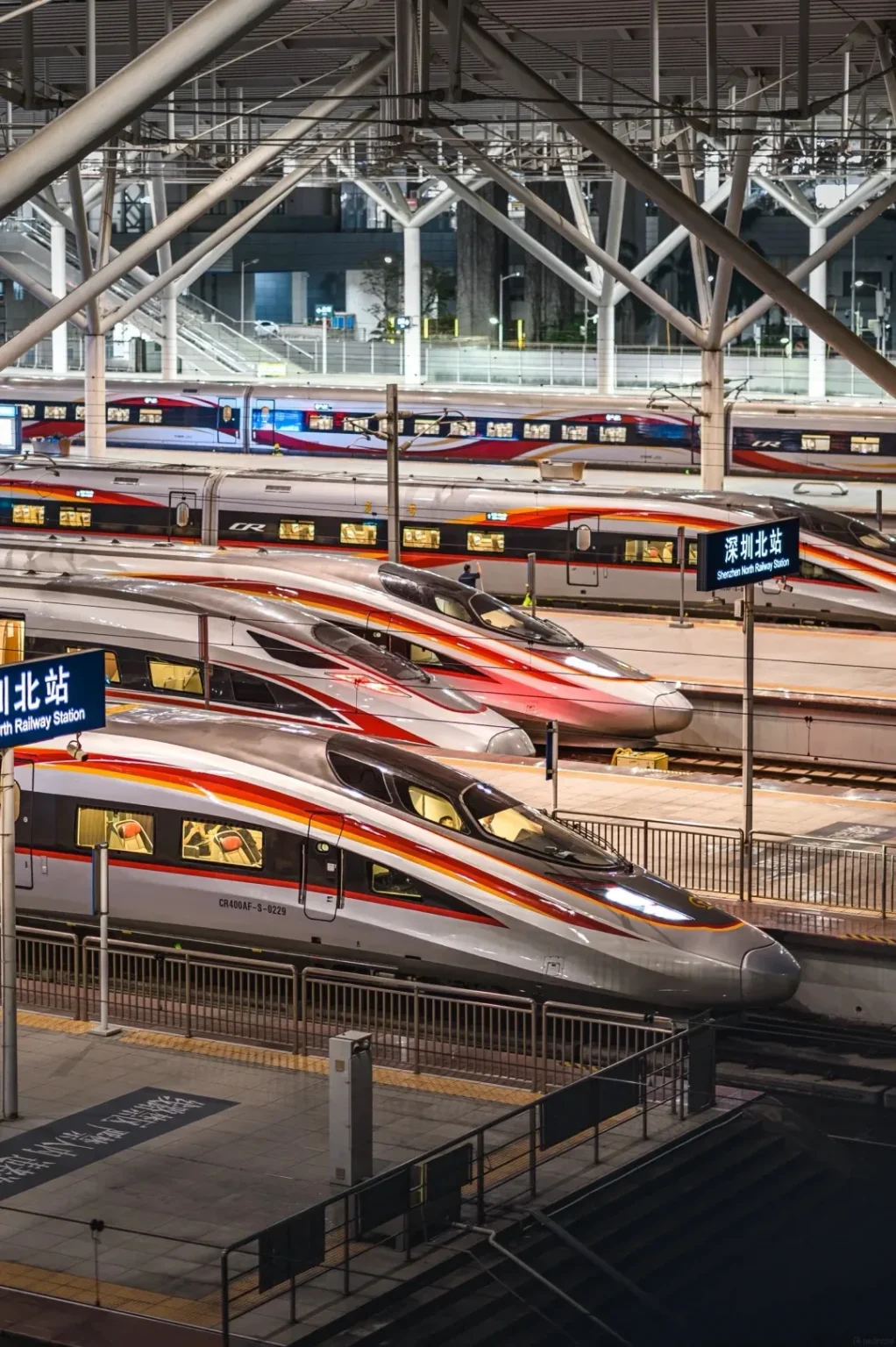 Several modern, silver and red high-speed trains are parked at a brightly lit indoor railway station. The trains have distinctive red and orange stripes along their sides. Overhead, a complex network of metal beams and cables forms the station's roof structure. A sign with Chinese characters and 'Shenzhen North Railway Station' in English is visible.