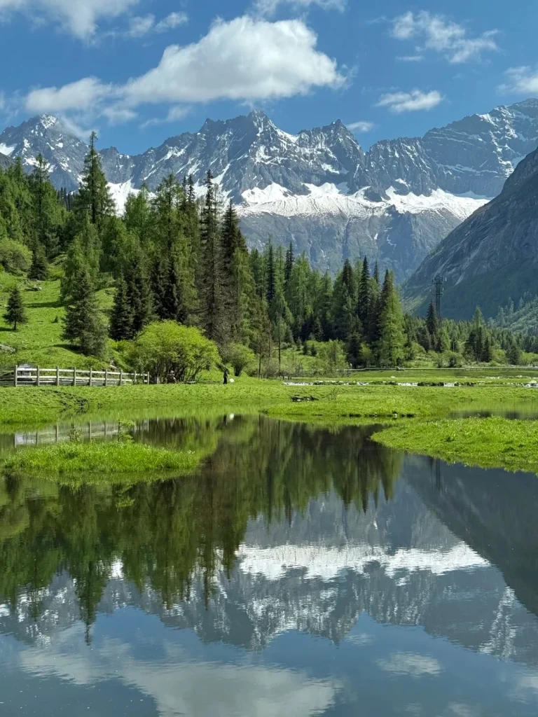 A serene lake reflects snow-capped mountains and lush green trees under a bright blue sky with white clouds. A wooden fence runs along a grassy hillside in the background, with a few people visible near trees.