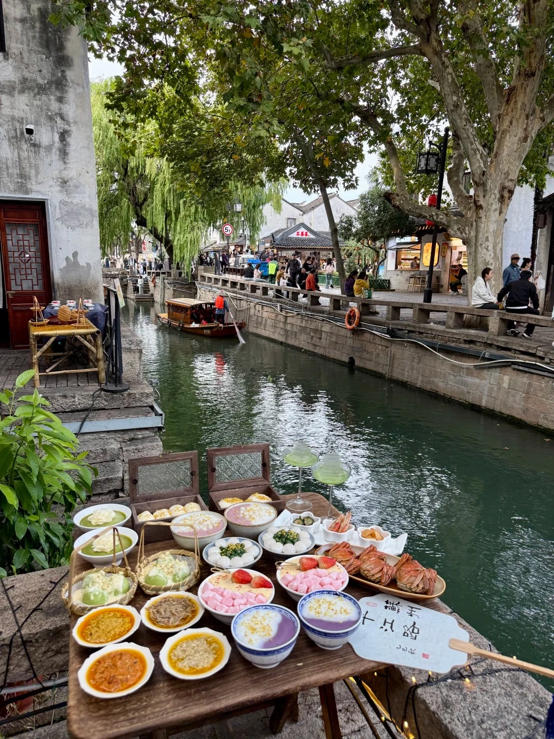 A table laden with various bowls of desserts and snacks sits on a stone ledge overlooking a canal in Suzhou. Dishes include purple sweet potato dessert, mochi, fruit-topped puddings, crab, shrimp, and other small appetizers. In the canal, a traditional wooden boat is being rowed by a person. Along the canal banks are ancient white-walled buildings with tiled roofs and lush green trees with weeping branches. People are visible walking and sitting along the canal's edge.