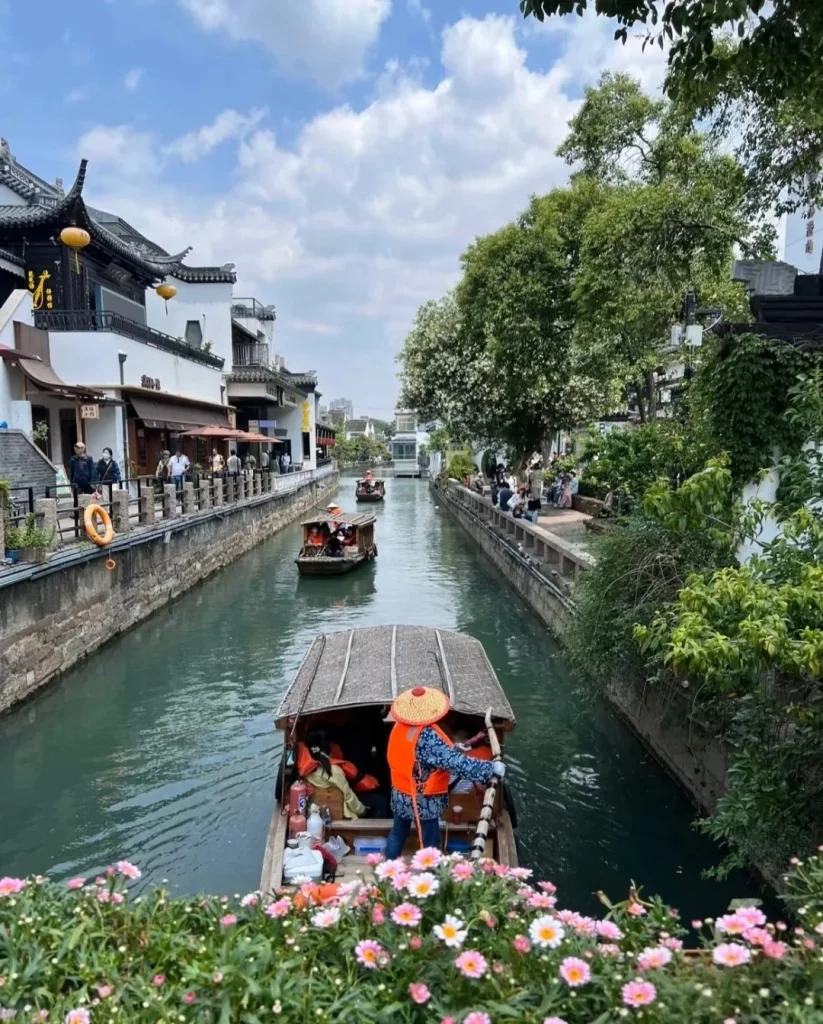 A traditional wooden boat with a thatched roof navigates a narrow canal lined with stone walls. A person wearing a conical straw hat and an orange life vest steers the boat with a long pole. Other boats with passengers are visible further down the canal, and traditional Chinese architecture with tiled roofs and lanterns lines the left bank. Lush green trees and vibrant pink and white flowers bloom along the canal's edge.