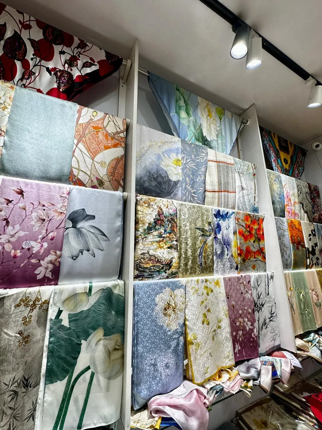 Shelves filled with folded silk scarves featuring various colorful patterns including floral motifs, paisleys, and geometric designs. Some scarves are draped over display shelves, while others are neatly stacked. The lighting illuminates the vibrant textures and intricate details of the silk.