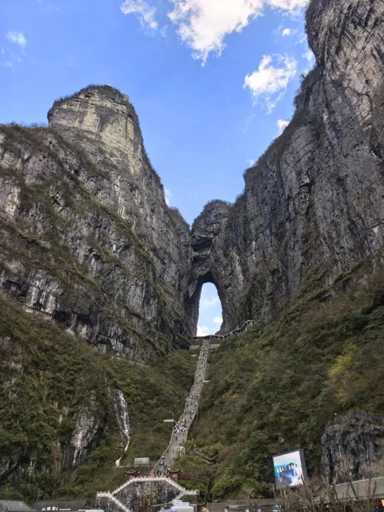 A view looking up at a natural archway in a massive rock formation, with a long, steep staircase filled with tiny tourists ascending towards it. Lush green vegetation covers the steep mountain slopes on either side of the staircase. A small waterfall cascades down the left side of the mountain.
