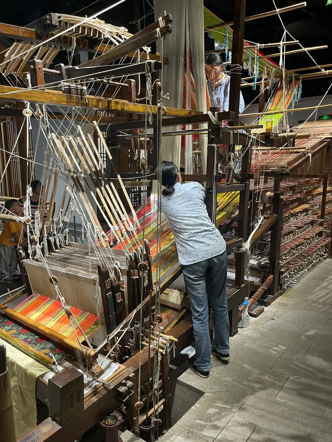 A person operating a large, intricate wooden loom with many threads and colorful fabric. Another person is visible above the loom, working with white threads. A smaller child stands to the left of the loom.