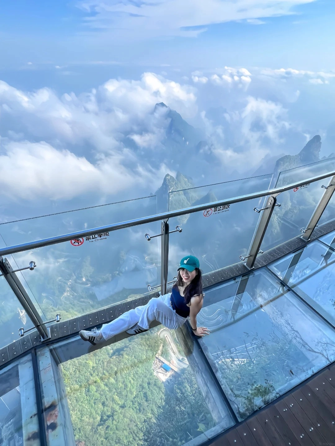 A young woman with a teal baseball cap, dark blue tank top, and white pants sits on a transparent glass skywalk. She is looking at the camera and smiling. The skywalk is elevated high above a mountainous landscape partially obscured by clouds. The view through the glass shows lush green mountains and winding roads below. A metal railing with warning signs is visible along the edge of the skywalk.