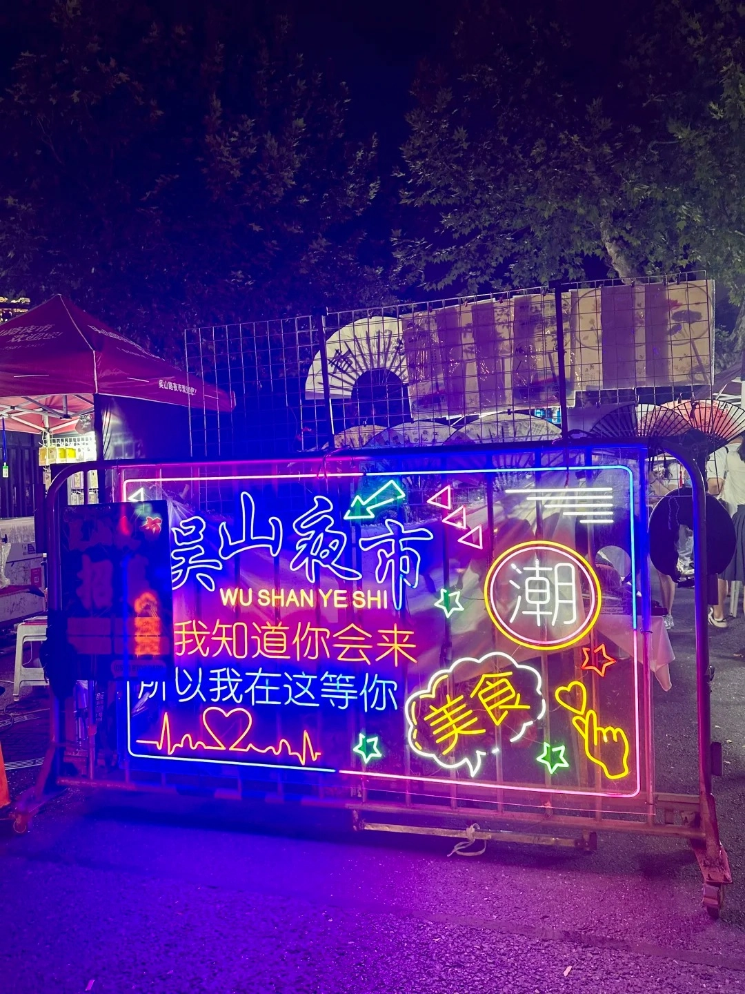 A bright neon sign for Wushan Night Market in Hangzhou at night, featuring Chinese characters that translate to 'Wushan Night Market,' 'I know you will come,' and 'So I'm waiting for you here.' The sign also includes neon illustrations of stars, hearts, a speech bubble with the word '美食' (meaning 'delicious food'), and a hand making a finger heart gesture. The sign is illuminated with vibrant blue, pink, yellow, and green neon lights.
