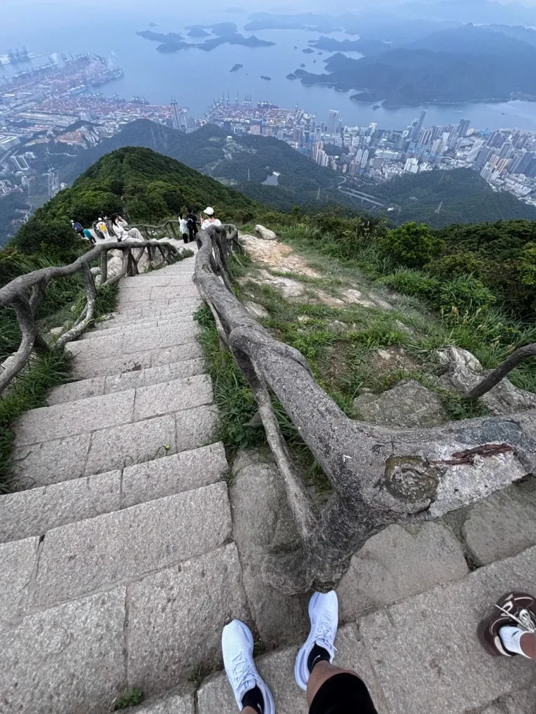 A person's feet wearing white sneakers stand at the bottom of a stone staircase that ascends a grassy, tree-lined mountain path. A wooden railing follows the path. In the background, a vast cityscape and harbor stretch out towards a hazy coastline and islands under an overcast sky.