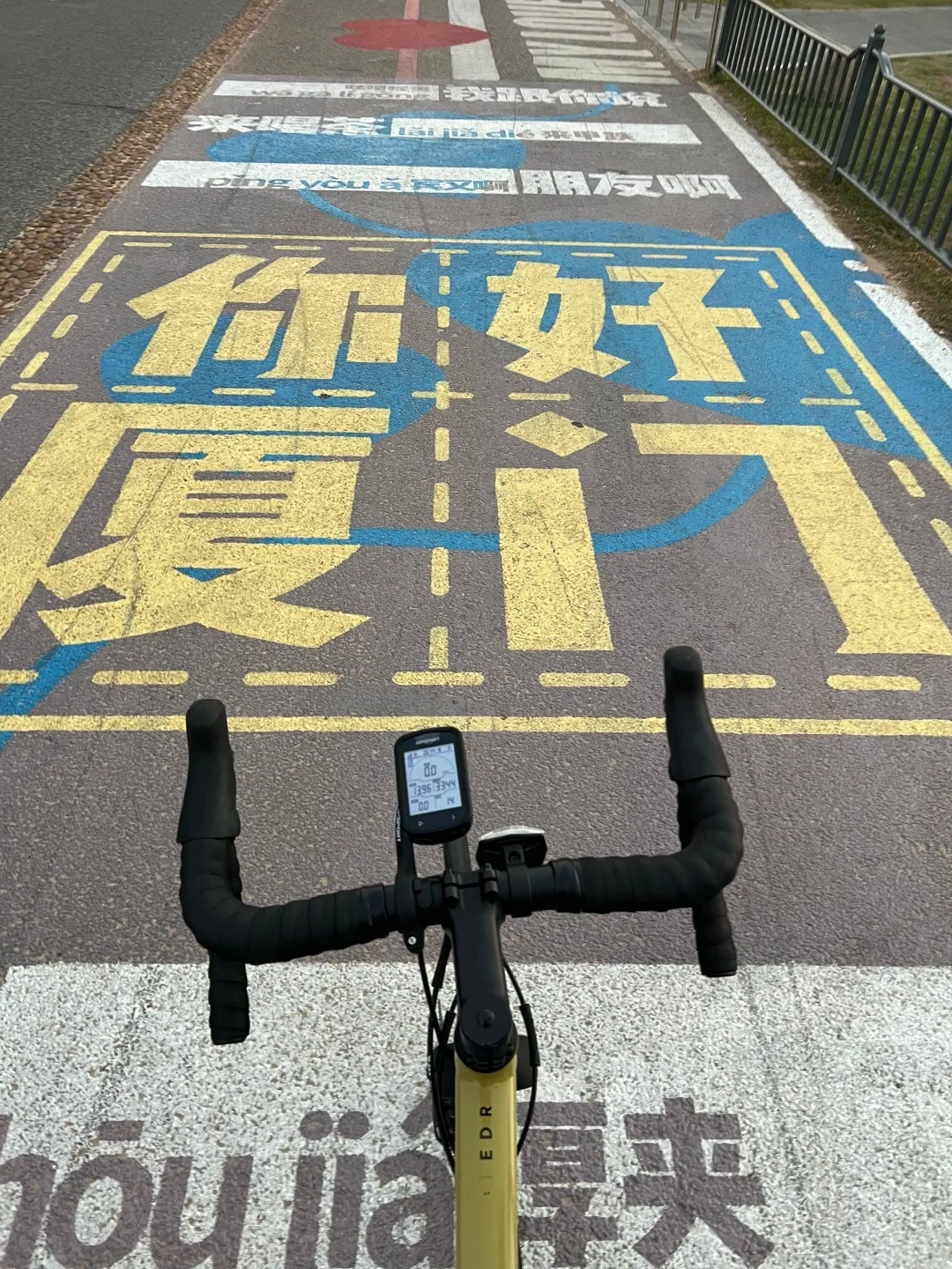A first-person view from a bicycle handlebar showing a yellow and blue painted path on asphalt with Chinese characters. The path has dashed lines and blocks of color, resembling a road lane. A bicycle computer is mounted on the handlebars.