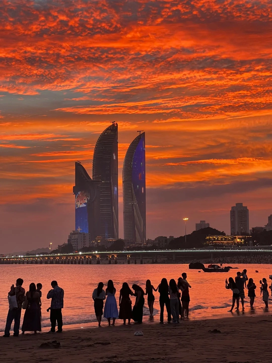 Silhouettes of people stand on a sandy beach and in shallow water at sunset. In the background, two modern skyscrapers with illuminated blue and red designs rise above a city skyline and a bridge lit with white lights. The sky is filled with dramatic, vibrant orange and red clouds.