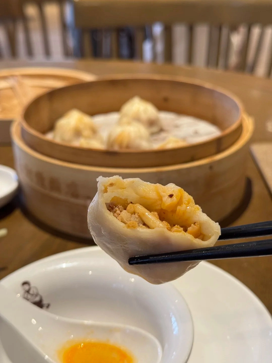 A close-up of a soupy xiaolongbao being held by black chopsticks over a white dish. The dumpling has a bite taken out of it, revealing its filling of minced meat and broth. In the blurred background, several more xiaolongbao are visible in a bamboo steamer.