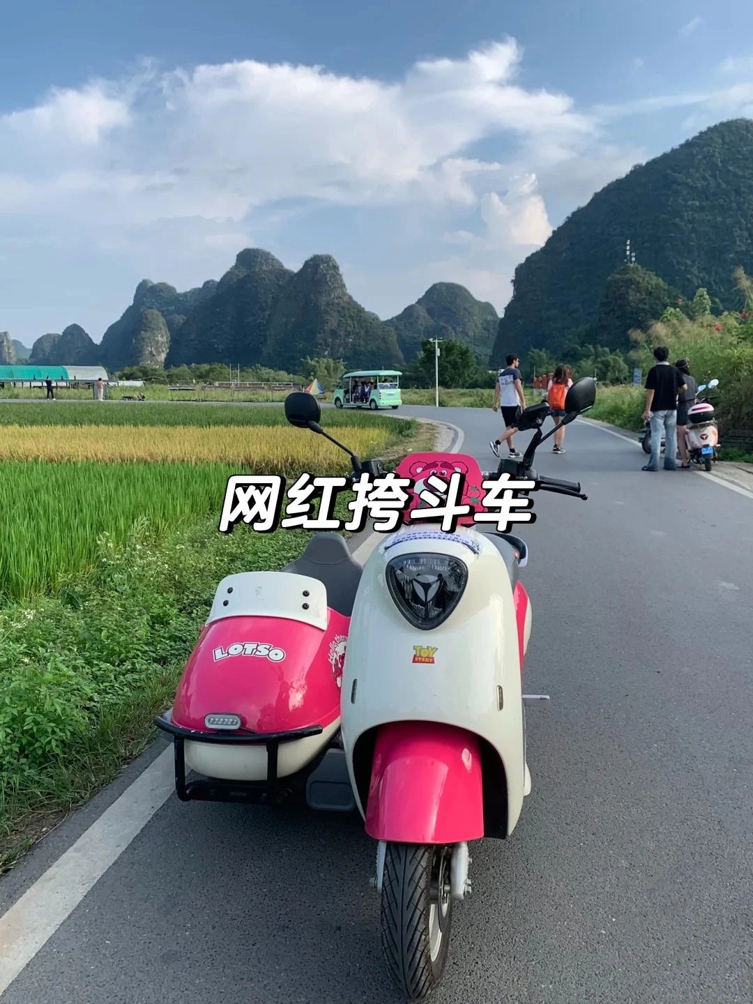 A pink and white electric scooter with a sidecar is parked on an asphalt road. In the background, lush green rice paddies stretch towards dramatic, tree-covered karst mountains under a cloudy blue sky. Several people are visible walking on the road and near a small green tourist vehicle in the distance.